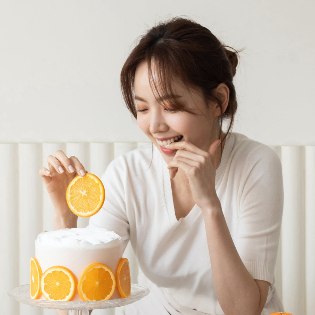 An Asian woman looking at a cake while holding a slice of orange
