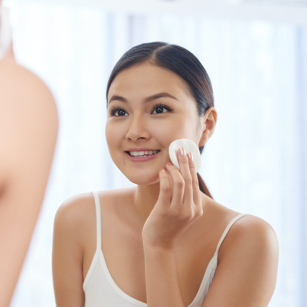 Smiling young woman applying toner on her face in front of mirror